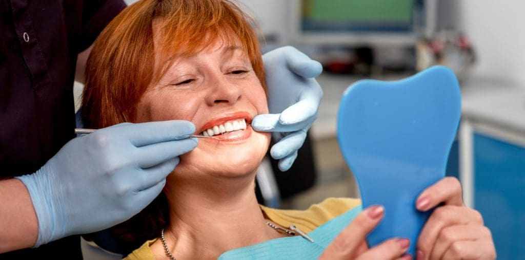 Smiling senior woman with new dental implants sitting in the dental office and looking at the mirror